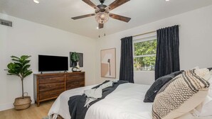 Une chambre à coucher avec un lit, une télévision, un ventilateur de plafond, une fenêtre avec des rideaux et une plante.