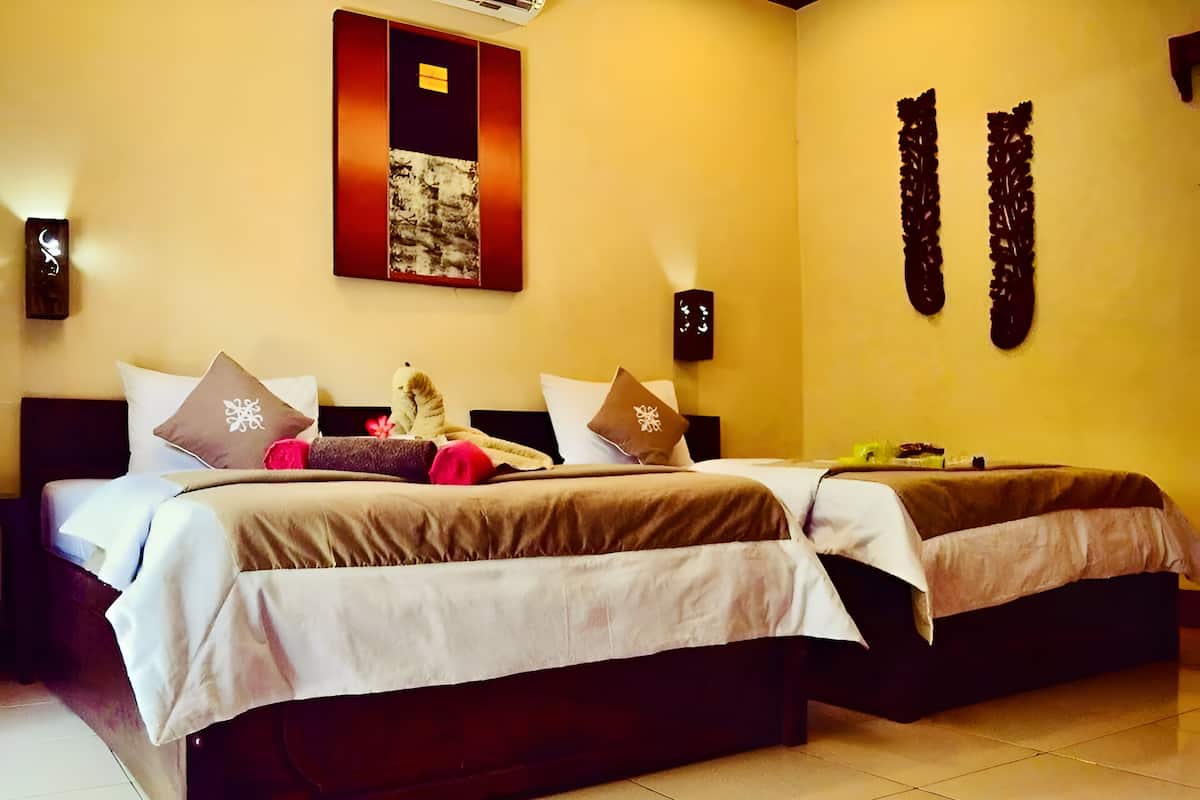 A hotel room with two beds, each with a brown bedspread and white linens. There are decorative pillows with snowflake patterns and a small statue on one bed. The room has a tiled floor and a wall-mounted air conditioning unit.