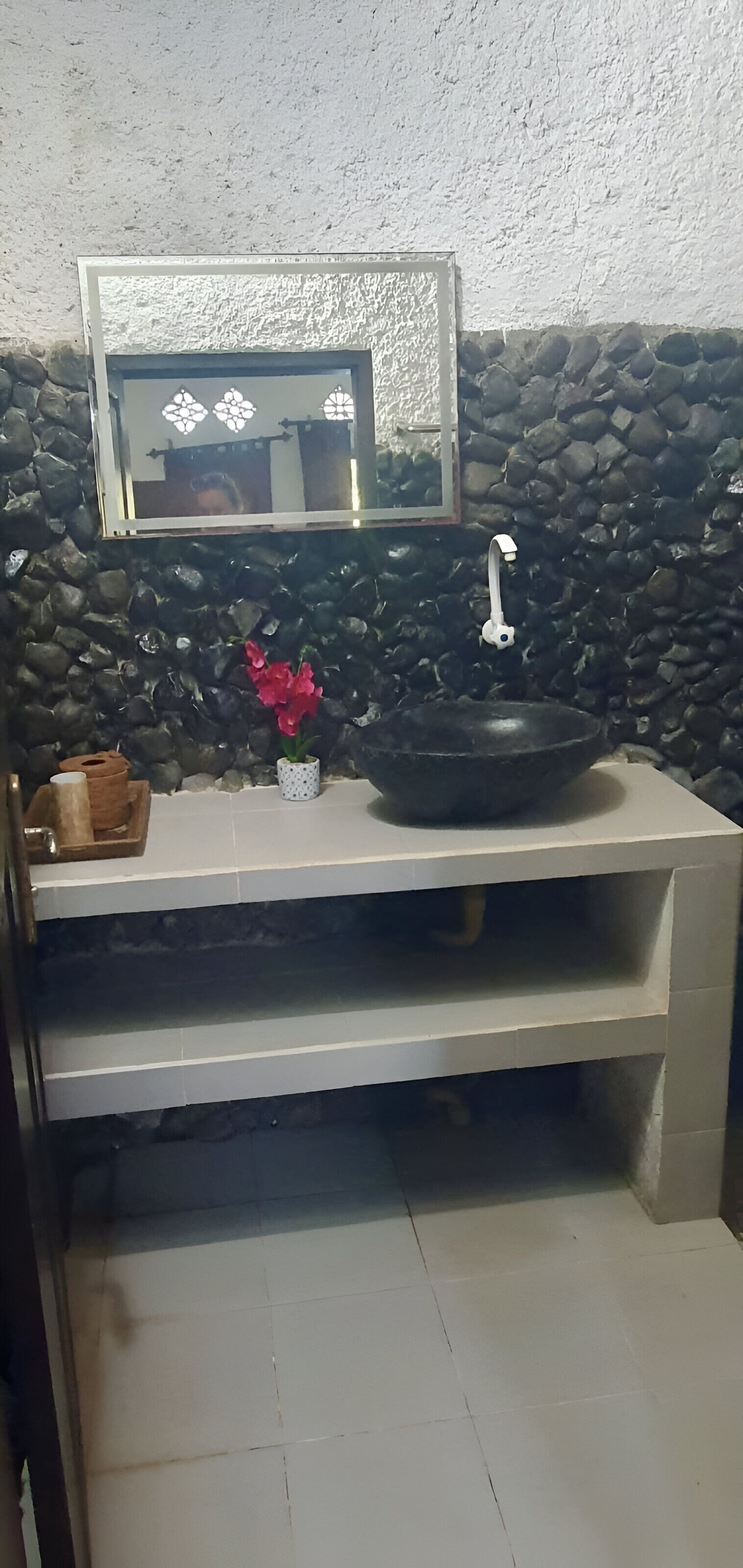A bathroom with a stone wall, a mirror, a black sink, and a small vase with flowers.