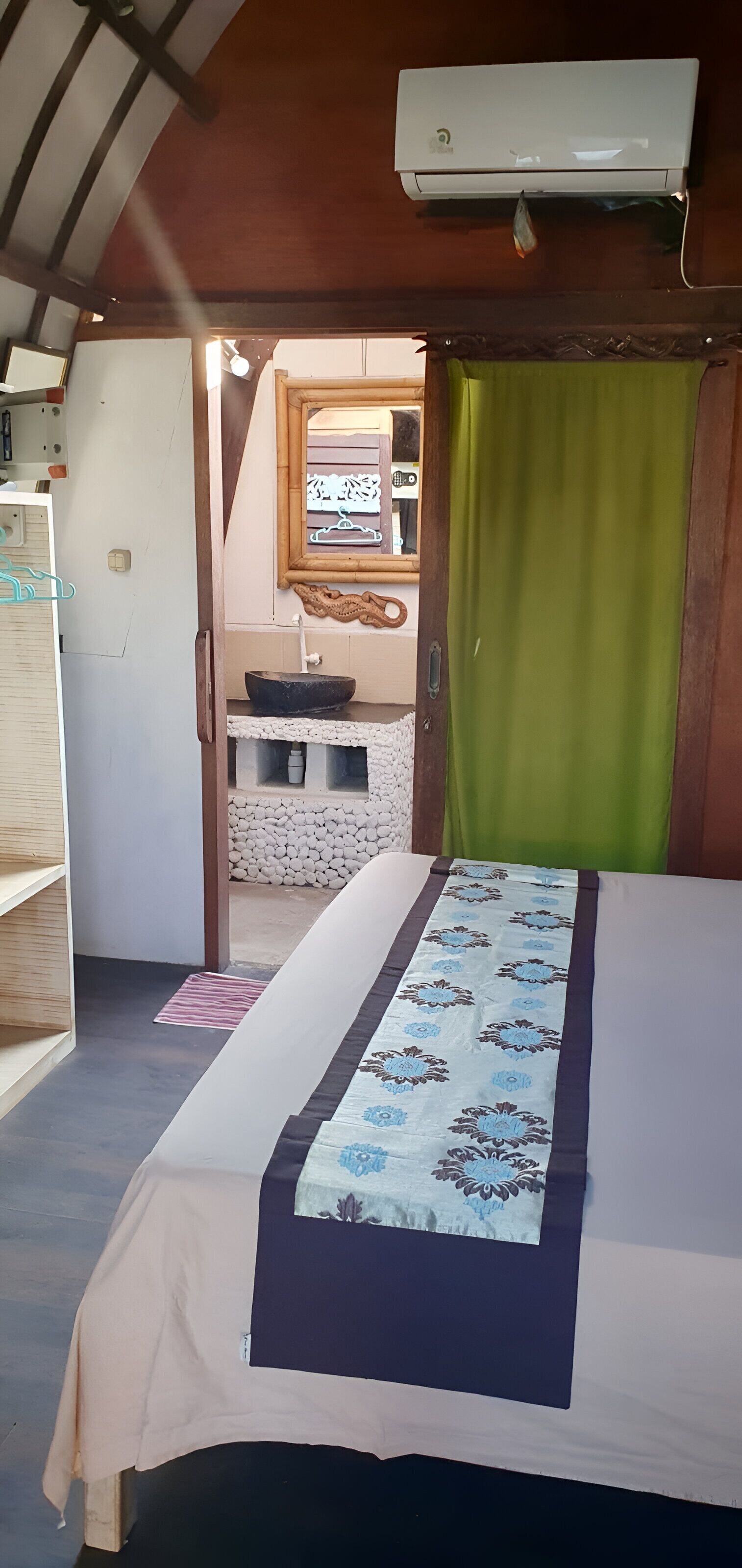 Un dormitorio con cama, cortina verde, puerta de madera, espejo y una pequeña cocina con pileta y estufa.