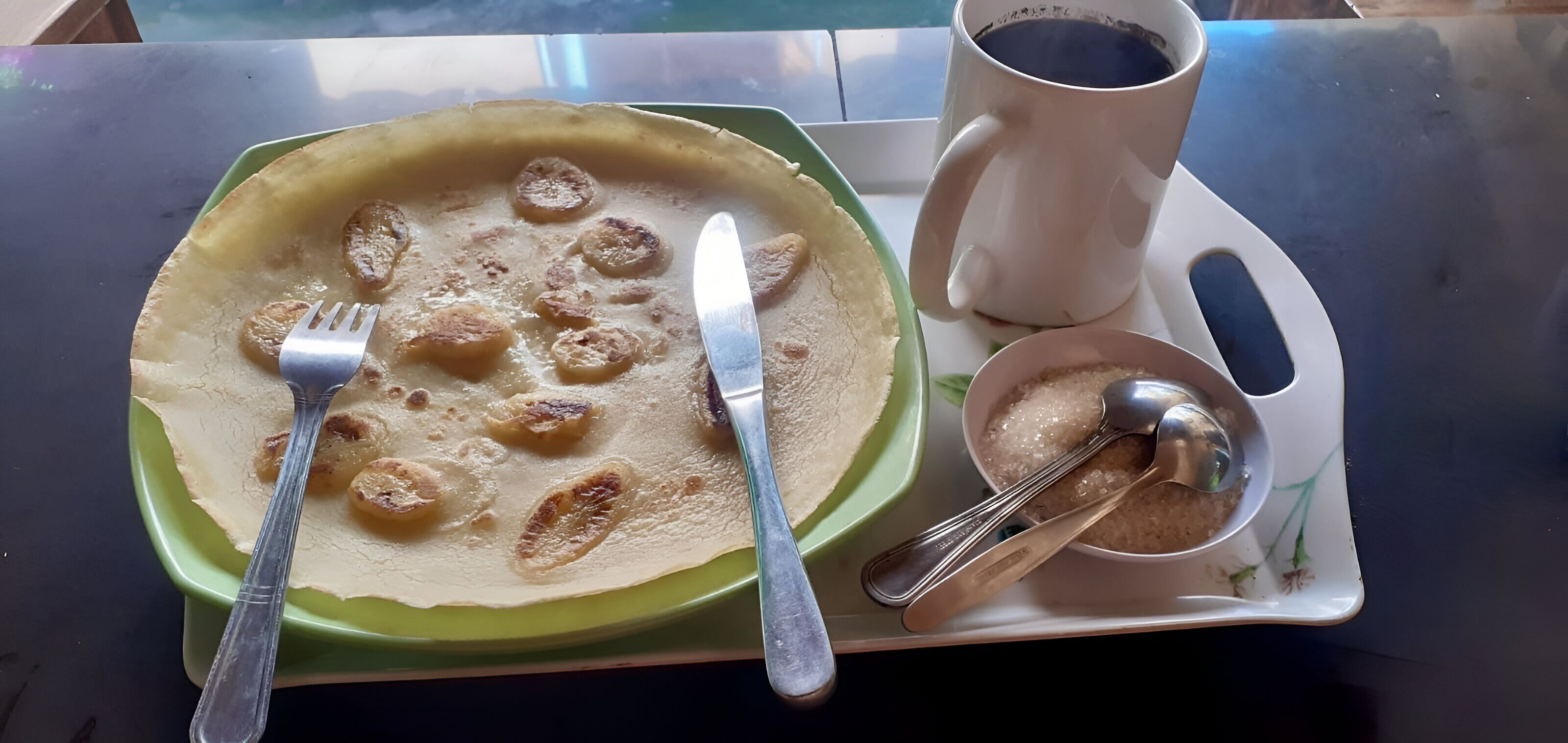 Un plateau repas avec une assiette verte contenant une crêpe aux fruits, une fourchette, un couteau, une tasse et une cuillère.