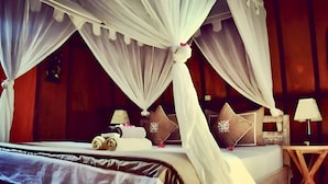 Una cama con dosel y cortina, con toallas enrolladas y almohadas decorativas, una mesita de noche con lámpara y un buró de madera.