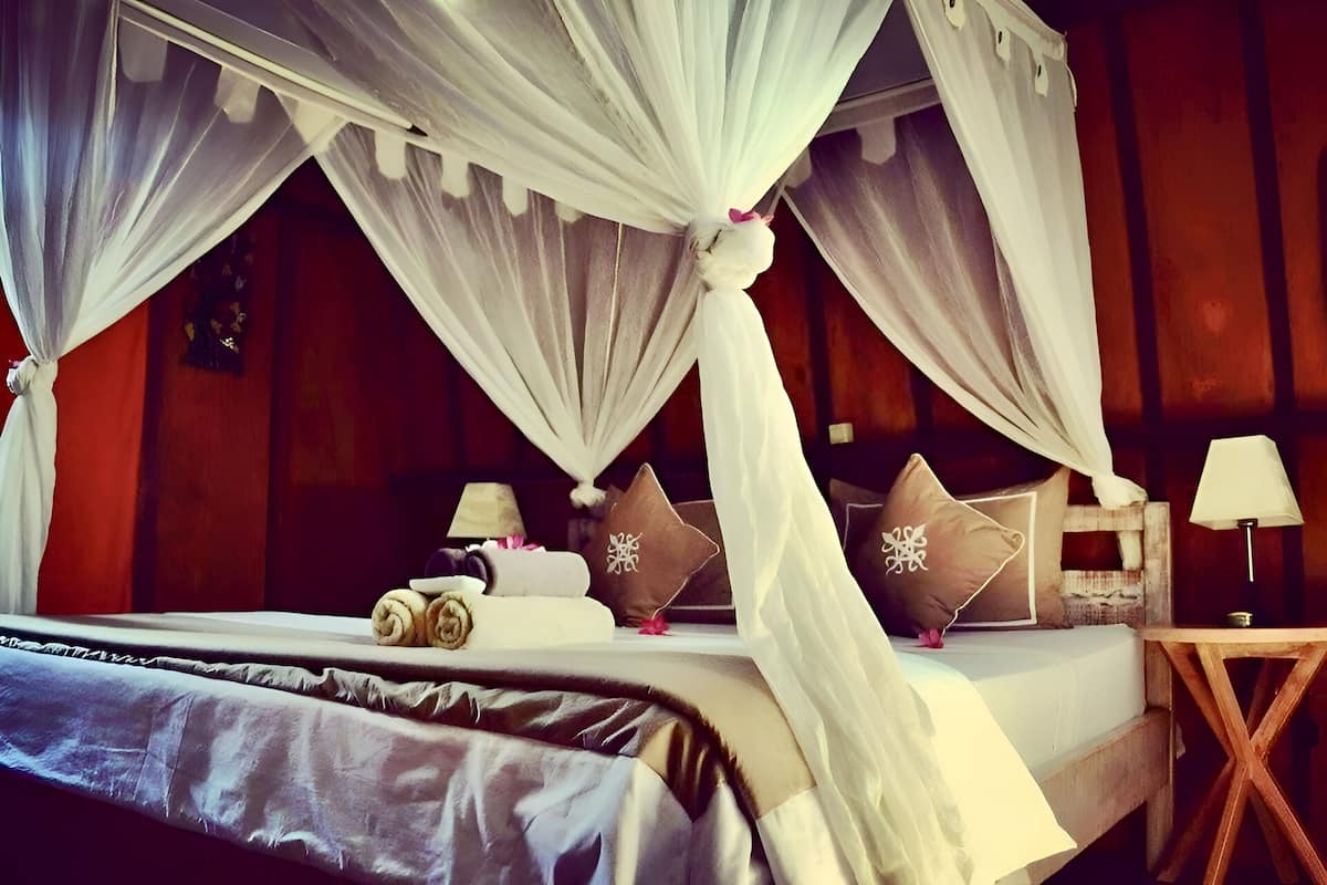 A four-poster canopy bed with rolled towels and decorative pillows, a bedside table with a lamp, and a wooden nightstand.