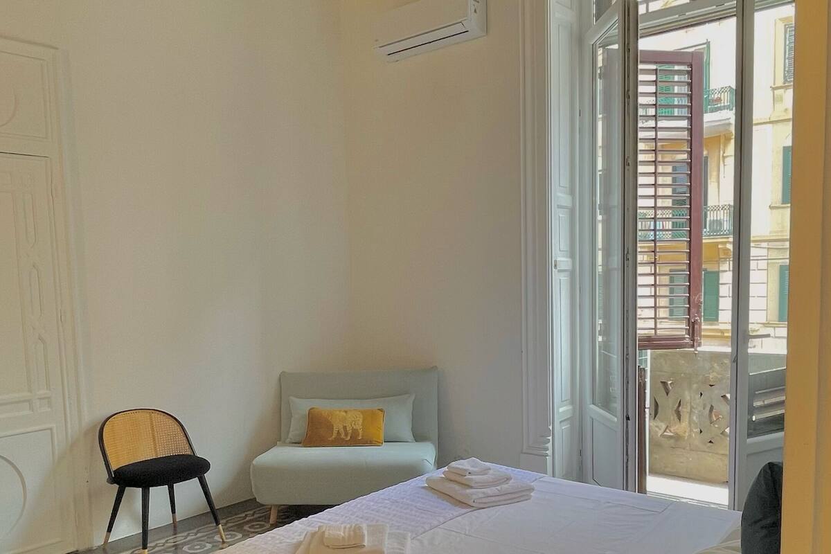 A hotel room with a bed, a chair, a window with a view of buildings, and a patterned floor.