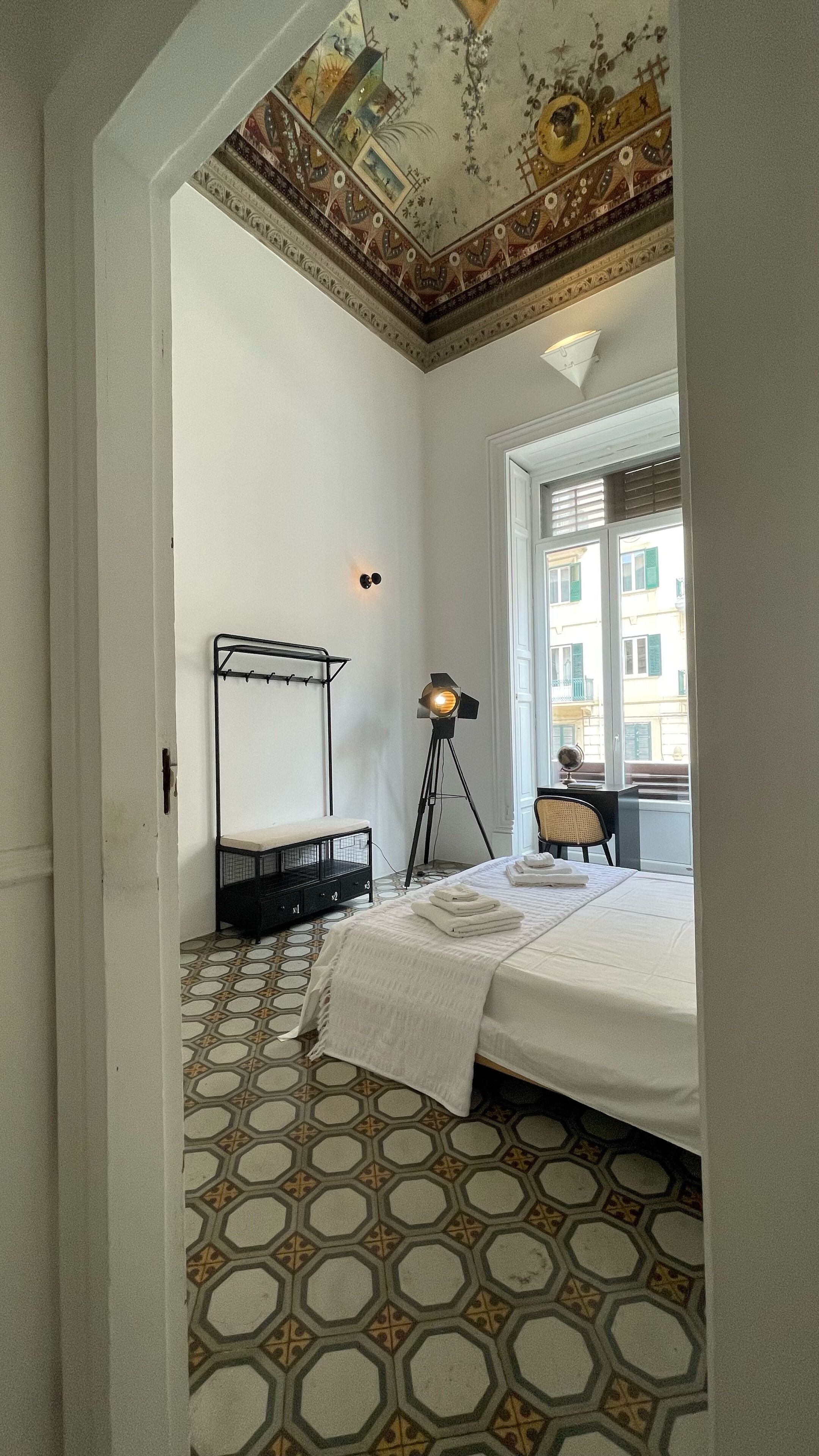 A bedroom with a bed, a floor lamp, a desk, and a view of buildings through a window.