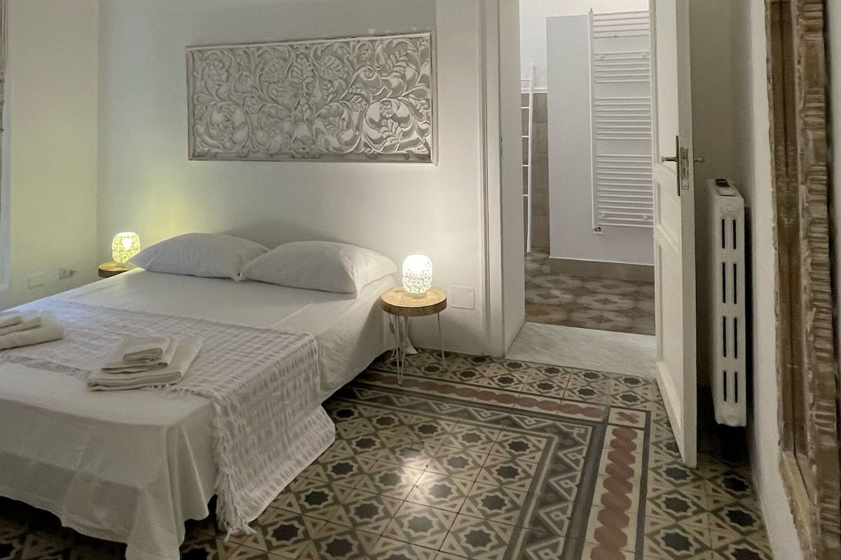 A bedroom with a bed, a nightstand, a wall-mounted artwork, patterned floor tiles, and a door leading to another room.