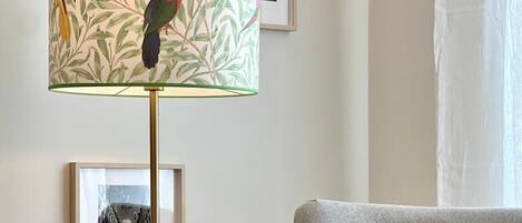Una habitación con un sillón gris, una lámpara de pie con estampado, una mesita redonda y cuadros en la pared.
