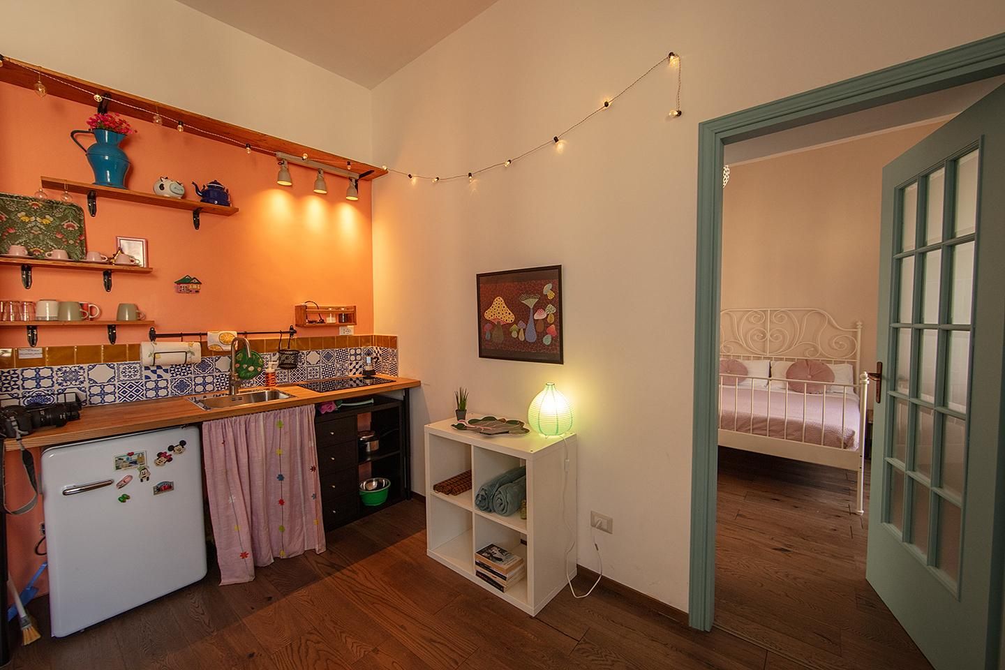 Eine kompakte Küche mit Kühlschrank, Spüle und Holz Arbeitsplatte. Ein kleiner weißer Tisch mit Lampe und einem Bild an der Wand. Ein Bett mit weißem Metallgestell ist durch eine offene Tür sichtbar.
