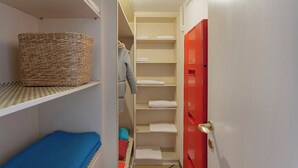 Un pasillo estrecho con una cesta de mimbre en una estantería, un colchón azul en el suelo, una chaqueta gris colgada y una puerta roja con cerradura.