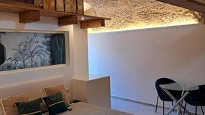 Ein modernes Zimmer mit Steindecke, einem Bett mit grün-beigen Kissen, einem Gemälde an der Wand und einem kleinen Tisch mit Stühlen.
