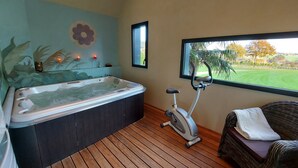 Una habitación con un jacuzzi, una bicicleta estática, una silla de mimbre y una ventana con vistas a un jardín y árboles.