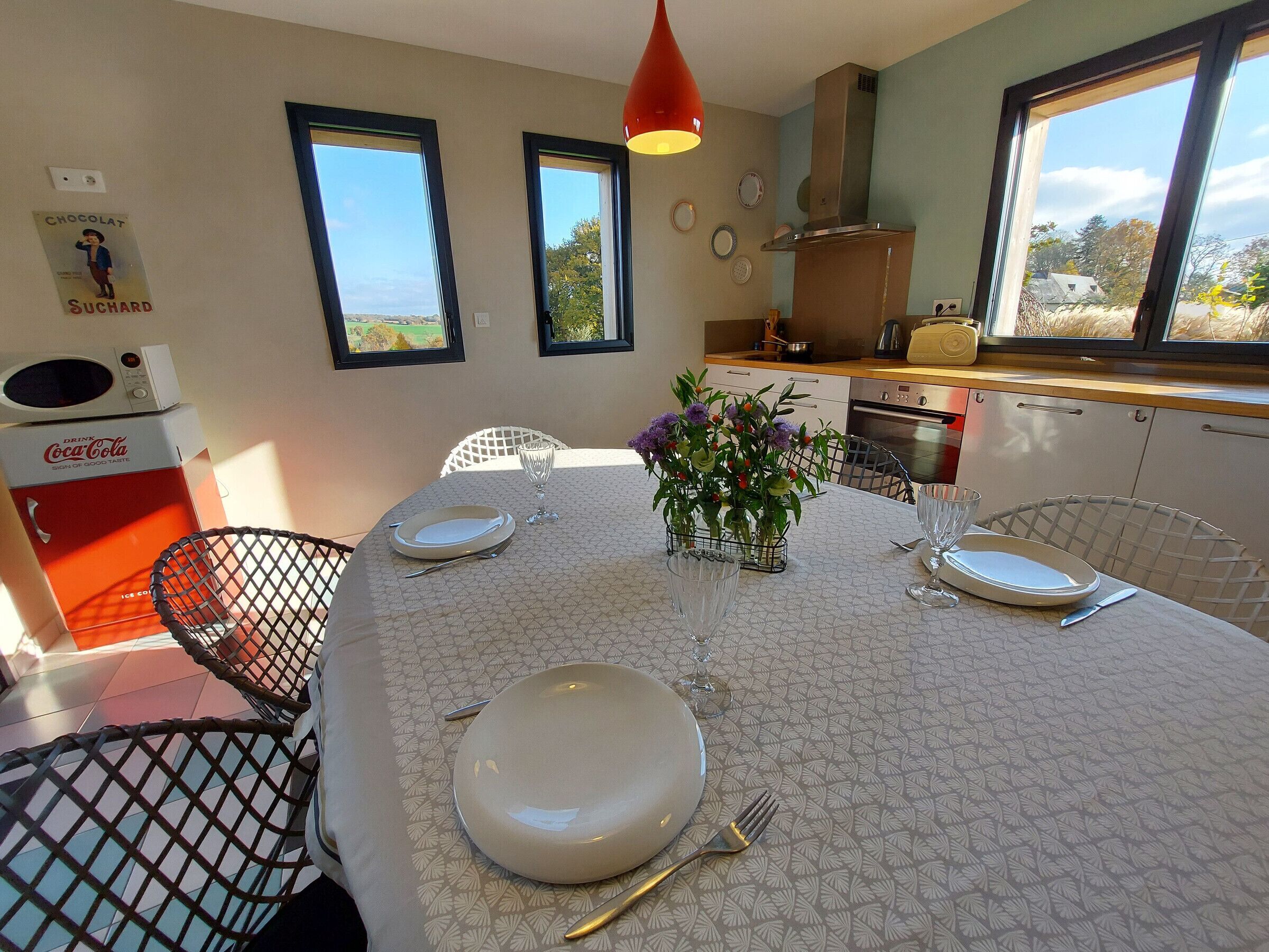 Un coin repas avec une table dressée pour quatre personnes, un vieux réfrigérateur Coca-Cola et une vue sur l’extérieur grâce à de grandes fenêtres.