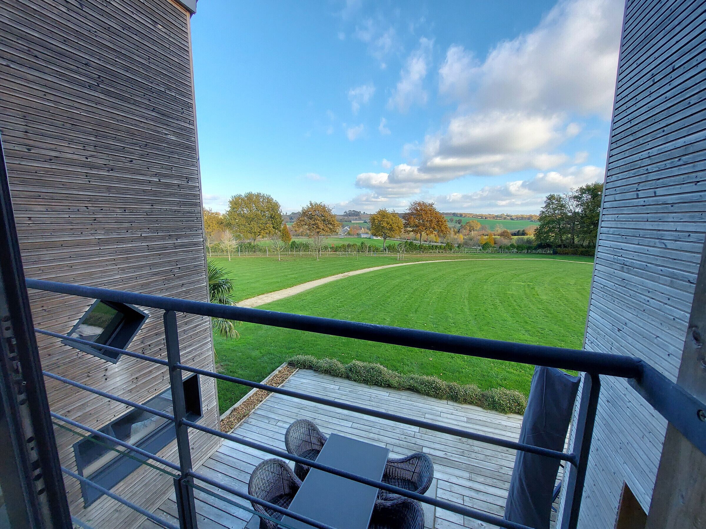Een balkon met uitzicht op een groen veld en bomen, een houten terras en een metalen balustrade.