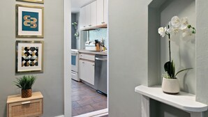 A modern kitchen with white cabinets, a built-in oven, and a sink. There is a potted plant on a wooden side table and a framed artwork on the wall.