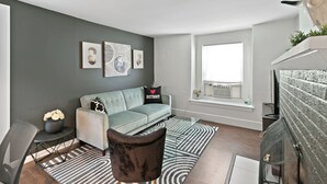 A modern living room with a grey sofa, a black armchair, and a glass coffee table. There are abstract wall art pieces and a window with a view.