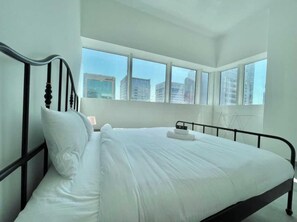 A modern hotel room with a large bed, white linens, and a city view through large windows. - Spacious Furnished Apartment in Palette Tower in Tourist Club Area (Abu Dhabi)