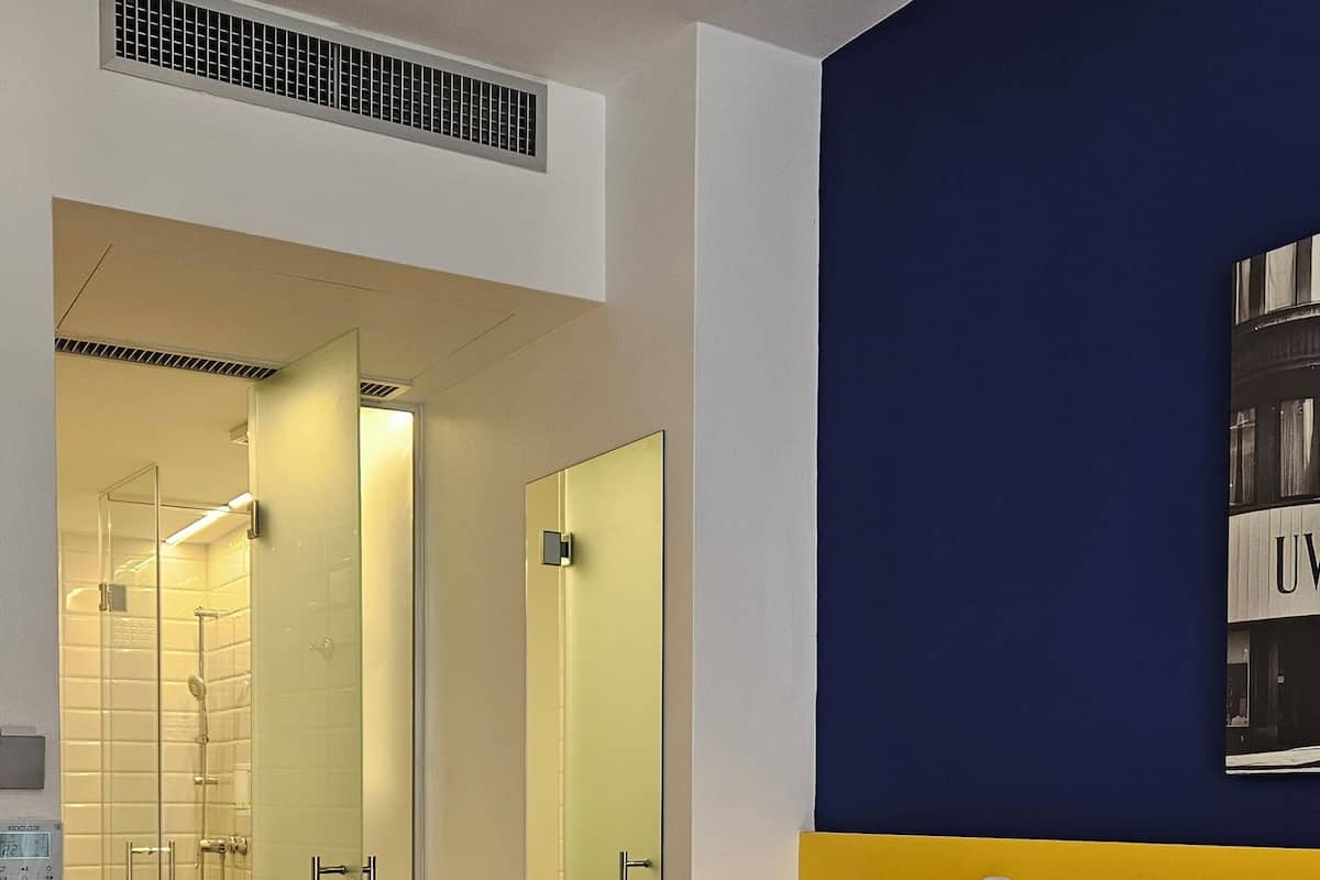 A hotel room with a bed, a bedside table, a nightstand, a mirror, a wall-mounted light, and a bathroom visible through an open door.