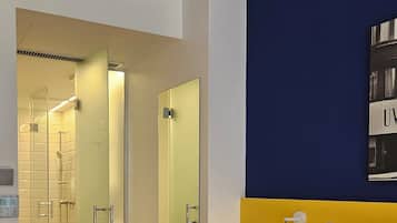 A hotel room with a bed, a bedside table, a nightstand, a mirror, a wall-mounted light, and a bathroom visible through an open door.
