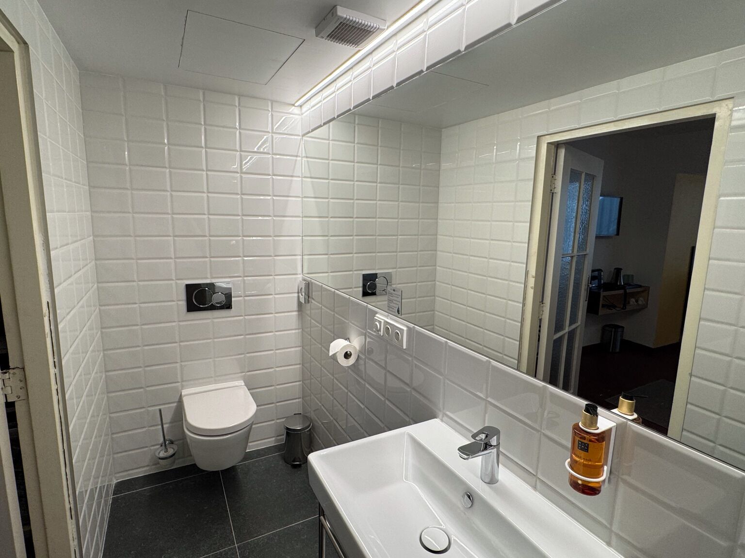 A bathroom with white tiled walls, a white toilet, a sink with a mirror, and a trash bin.