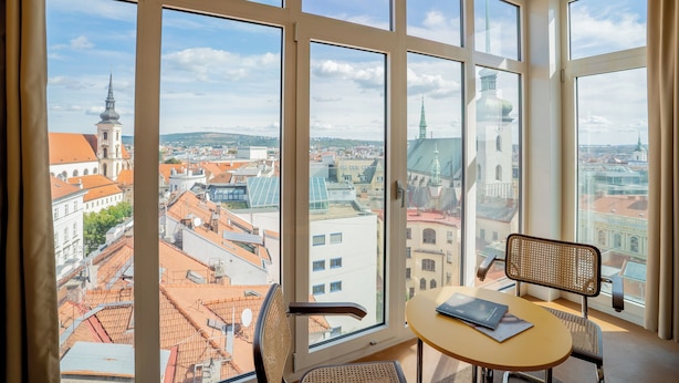 A room with large windows offering a cityscape view, a round table, and a wicker chair. - Hotel Avion - National Cultural Monument - ICONIC HOUSES (Brno)