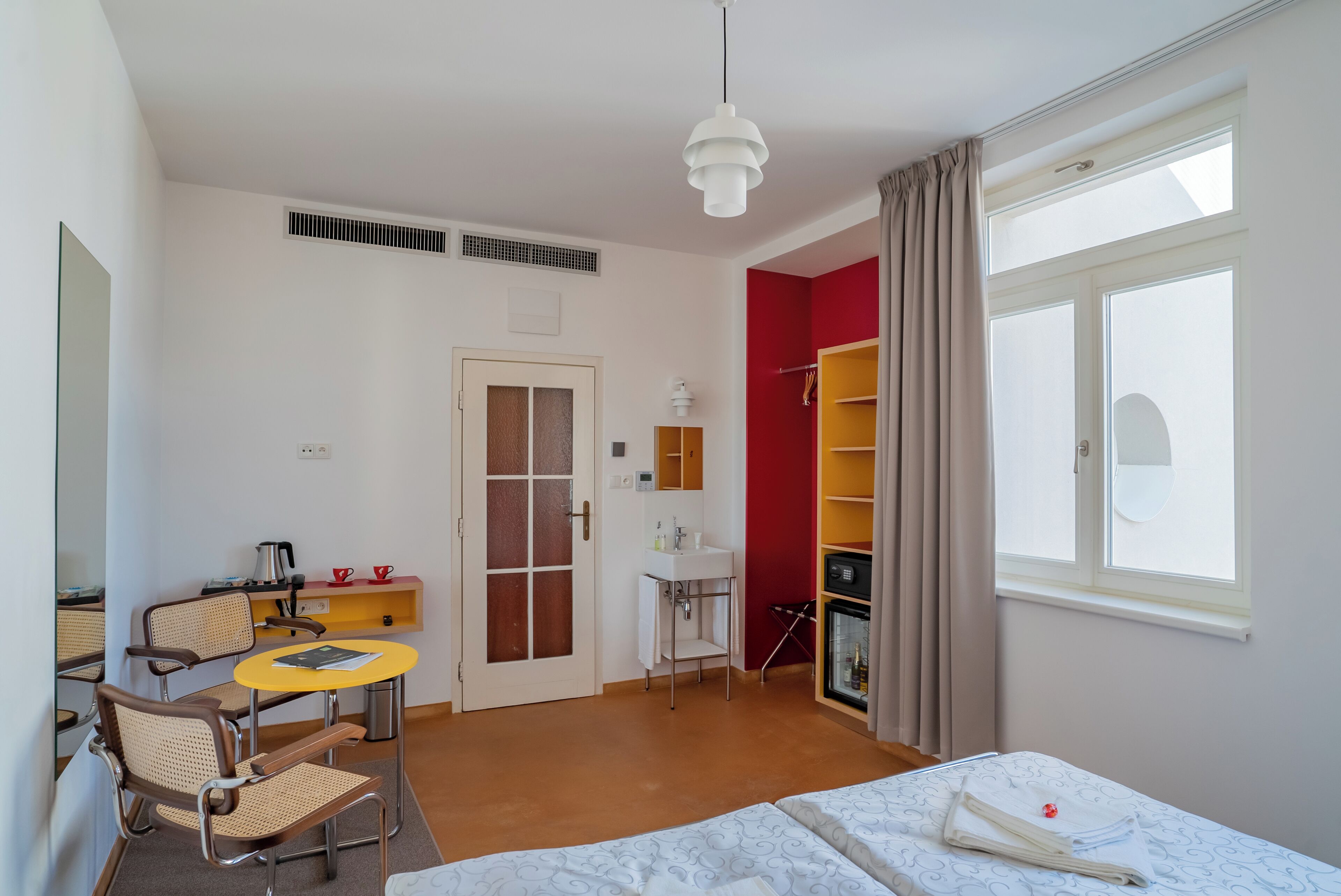 A hotel room with a bed, a desk with a chair, a small table, a wardrobe, and a window with curtains.