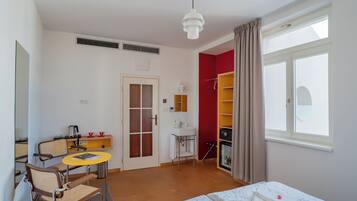 A hotel room with a bed, a desk with a chair, a small table, a wardrobe, and a window with curtains.