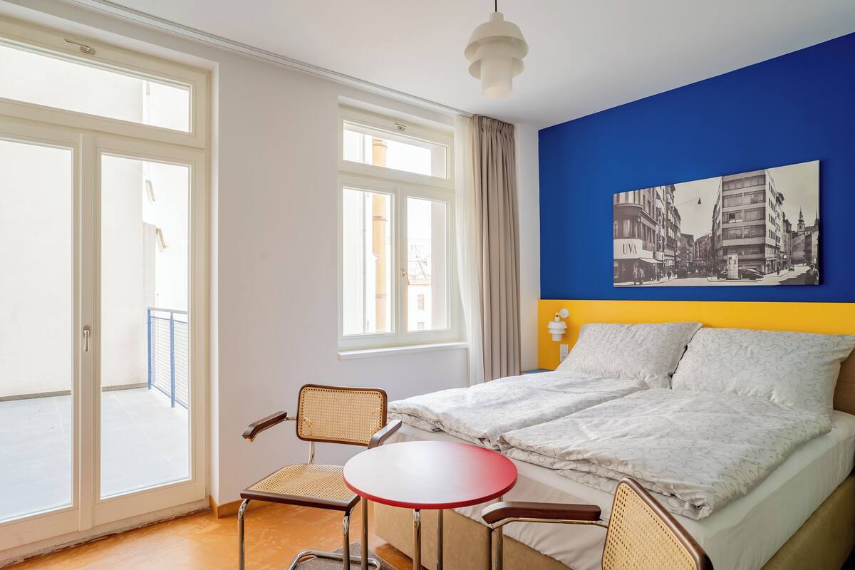 A hotel room with a bed, two chairs, a small table, a window with curtains, and a wall-mounted picture.