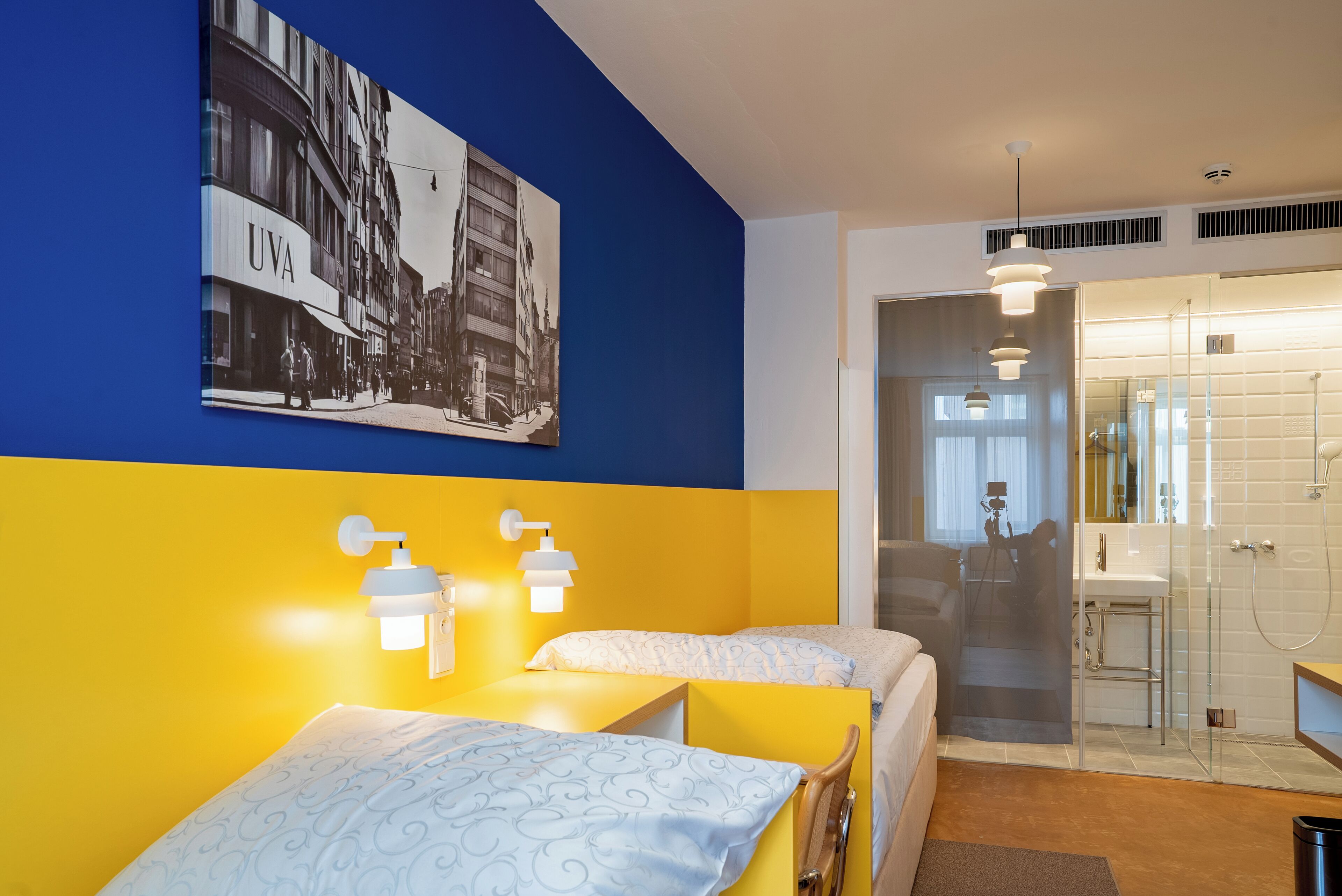 A hotel room with two beds, a bathroom with a glass shower, and a wall-mounted picture of a cityscape.