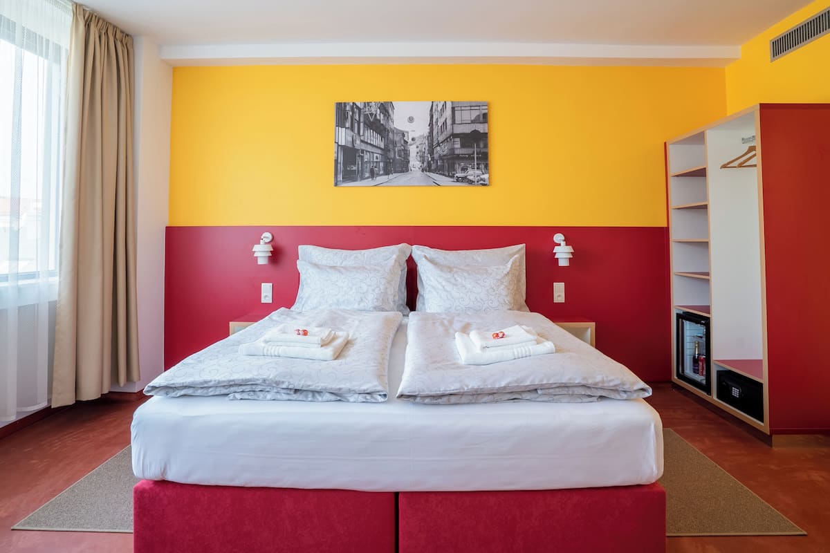 A double bed with white linens and pillows, a red and yellow wall, a black and white cityscape painting, a wardrobe, and a minibar.