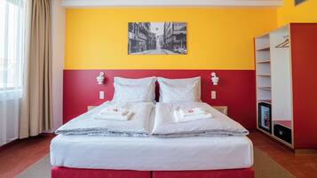 Una cama doble con ropa de cama y almohadas blancas, una pared de color rojo y amarillo, un cuadro de un paisaje urbano en blanco y negro, un armario y un minibar.
