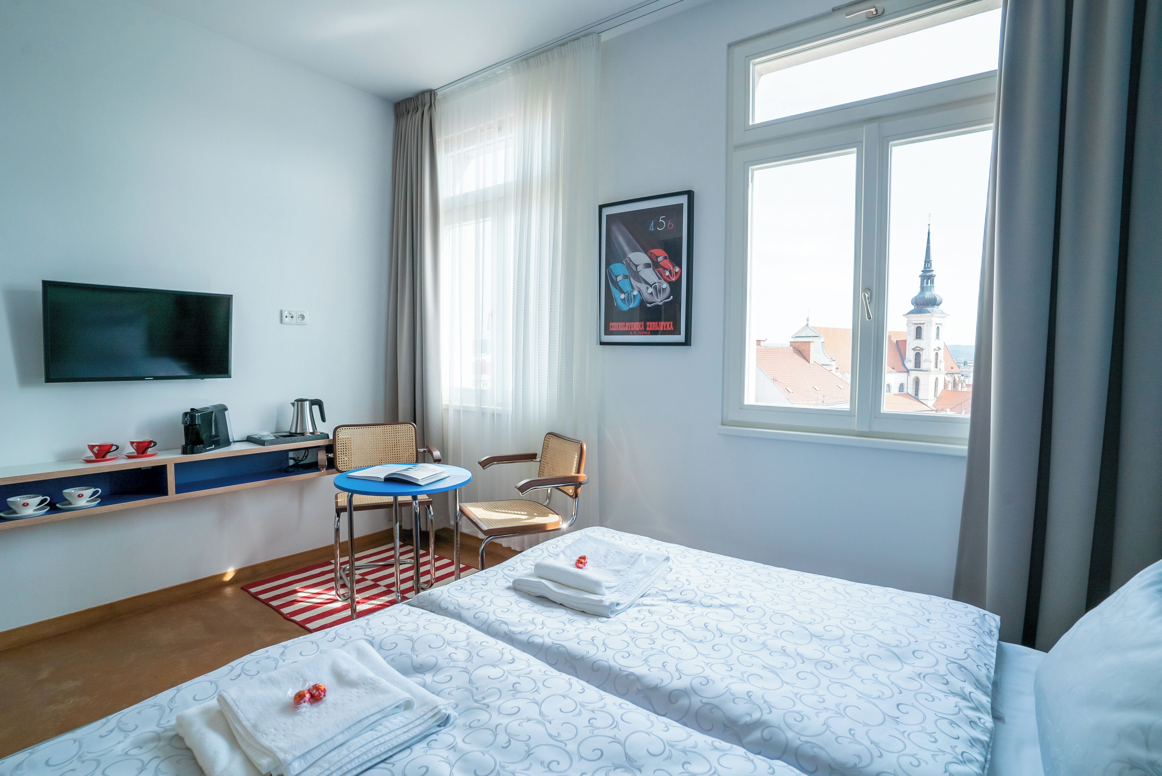 A hotel room with a bed, a TV, a desk, and a chair. There is a window with a view of buildings outside.