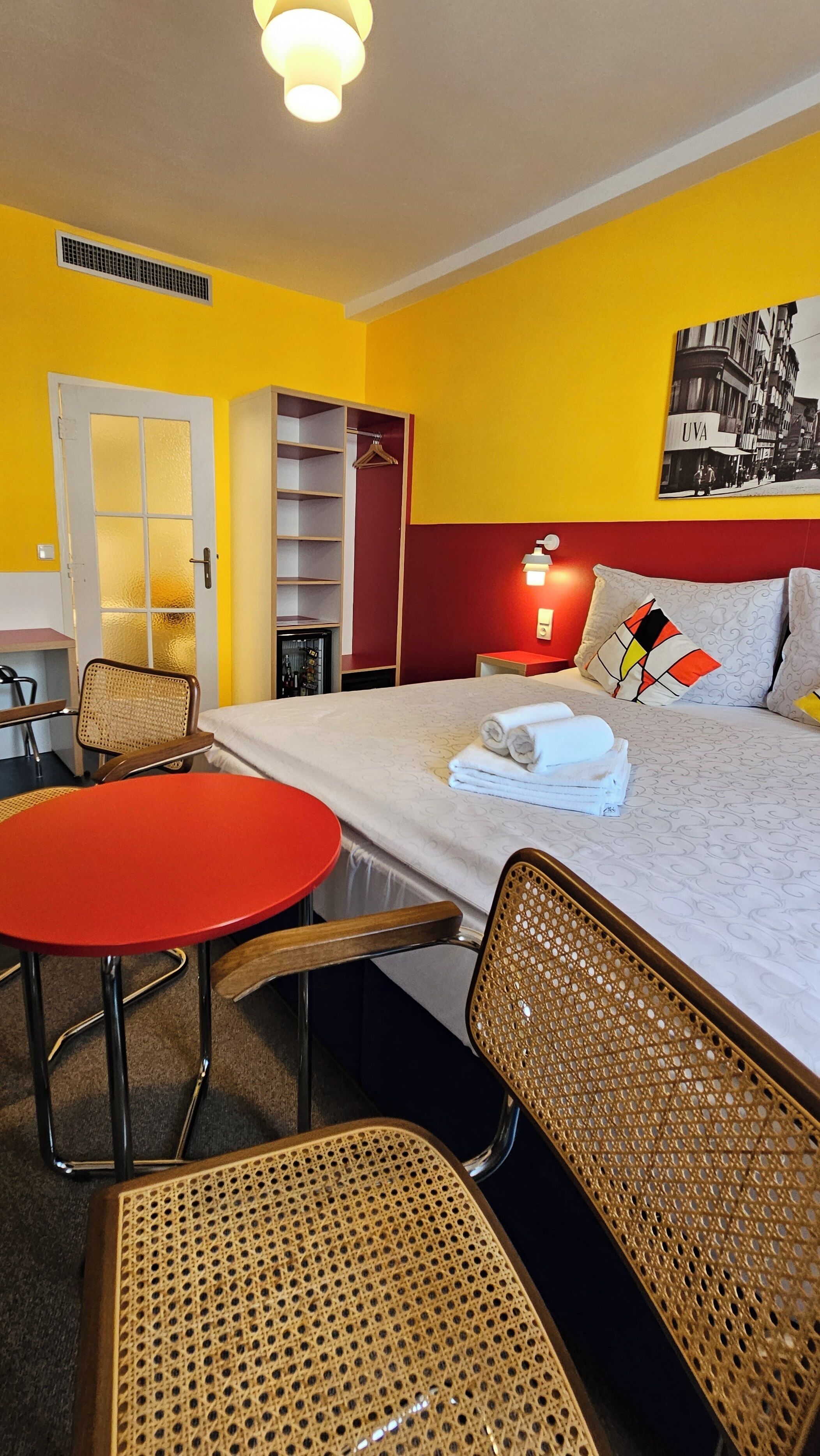 A hotel room with a bed, a red table, wicker chairs, a wardrobe, and a wall-mounted lamp.