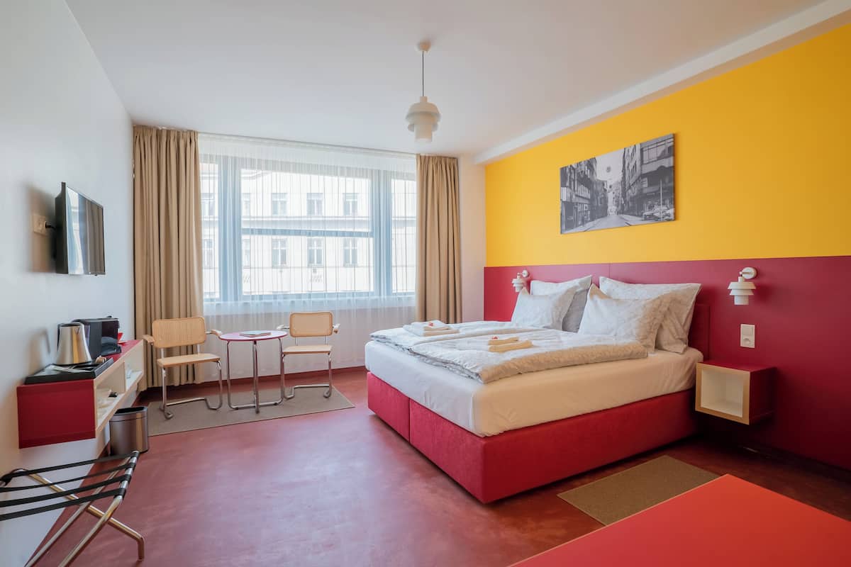 A modern hotel room with a red and yellow color scheme, a bed with white linens, a desk with a chair, a small table, and a window with curtains.