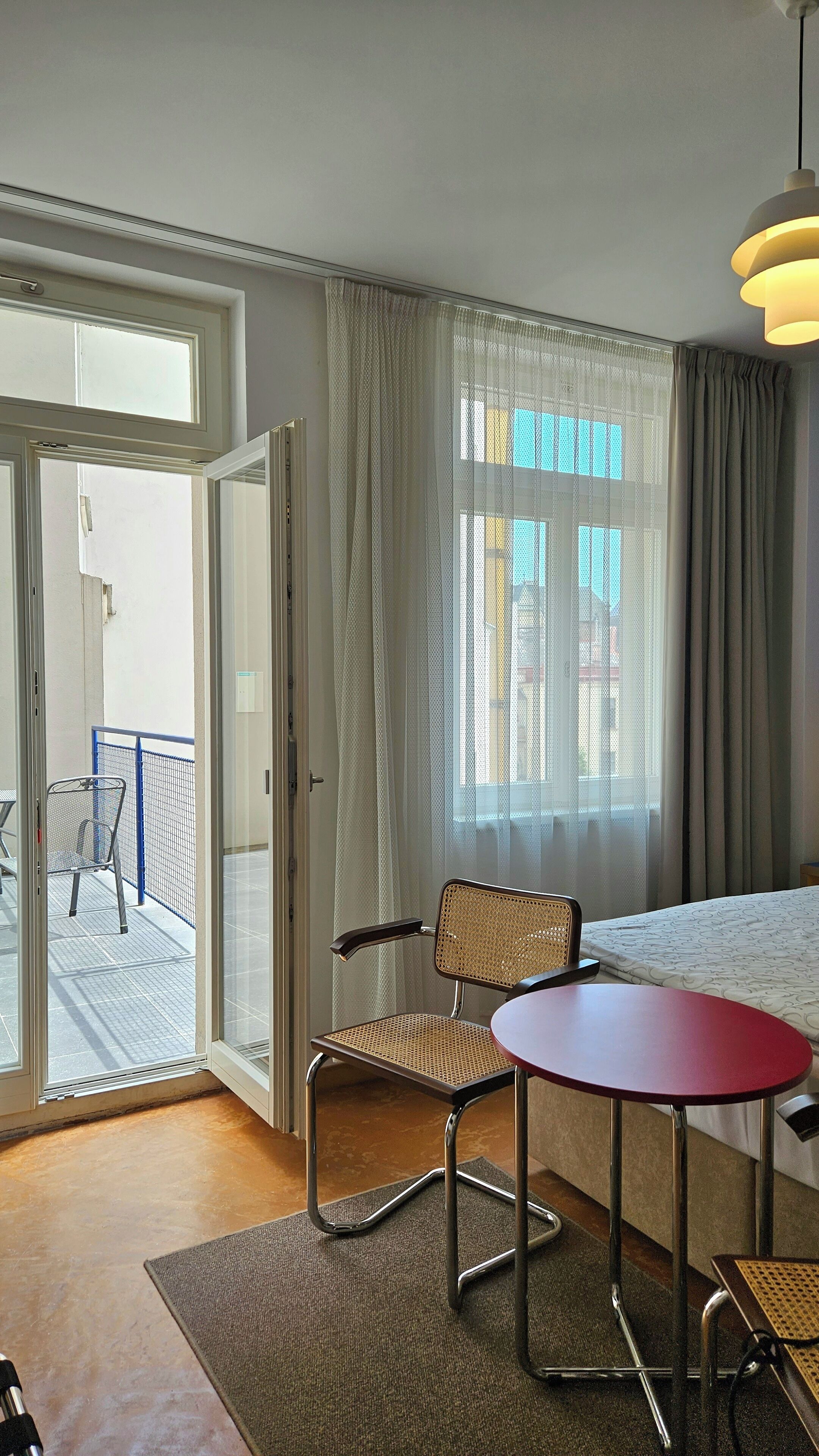 A hotel room with a bed, a chair, a small table, a window with curtains, and a view of the outside balcony.