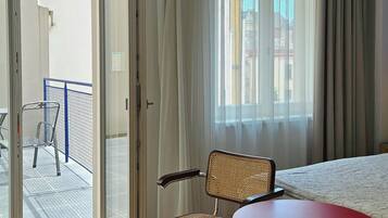 A hotel room with a bed, a chair, a small table, a window with curtains, and a view of the outside balcony.