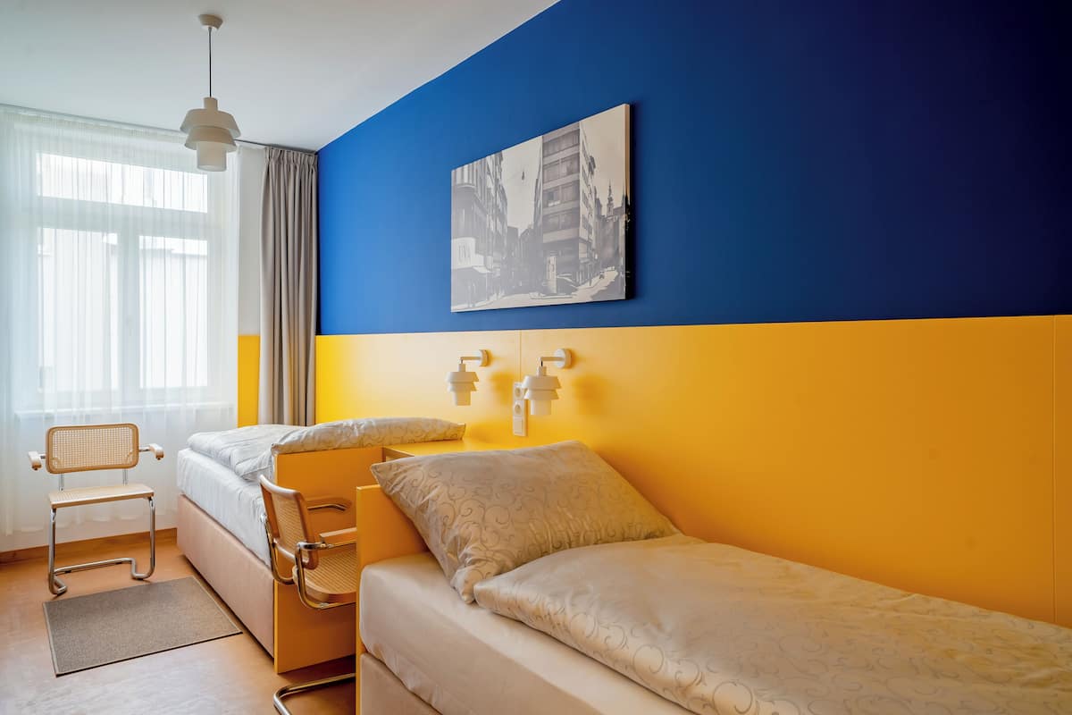 A room with two beds, a chair, a painting on the wall, and a window with curtains.