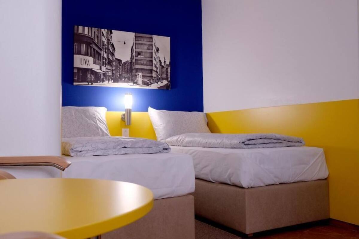 A hotel room with two beds, a small table, and a wall-mounted light.
