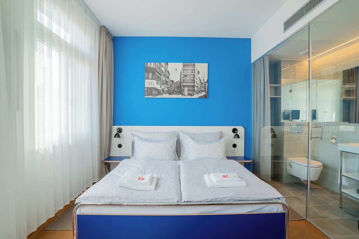 A modern hotel room with a large bed, a cityscape painting on the wall, and a glass shower enclosure.