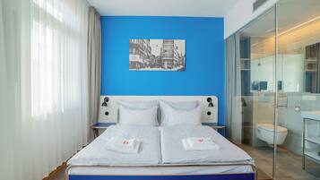 A modern hotel room with a large bed, a cityscape painting on the wall, and a glass shower enclosure.
