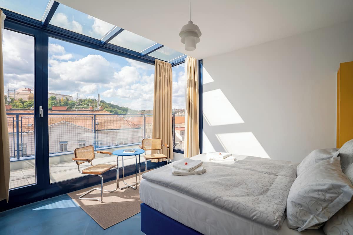 A modern hotel room with a large balcony, a bed with neatly made linens, a small table and chair, and a view of the cityscape.