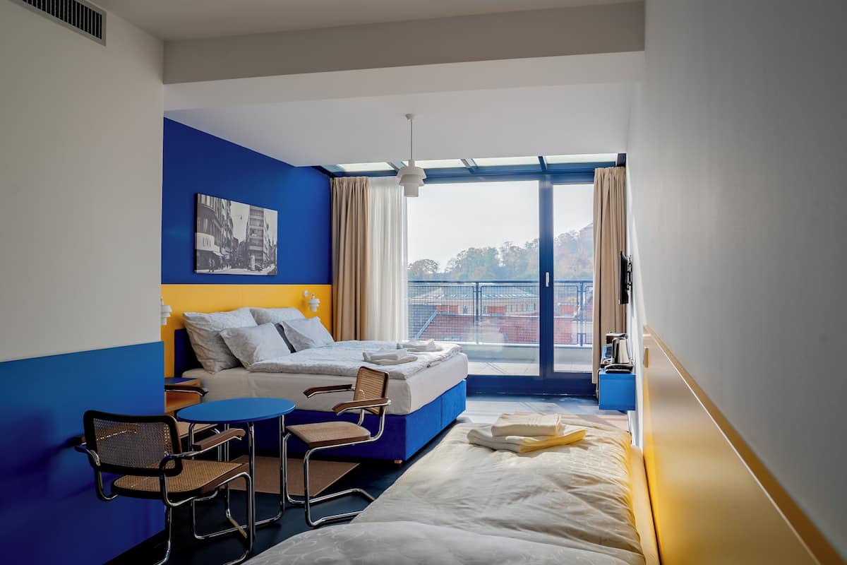 A hotel room with a bed, a small table, a chair, and a window with a view of buildings.