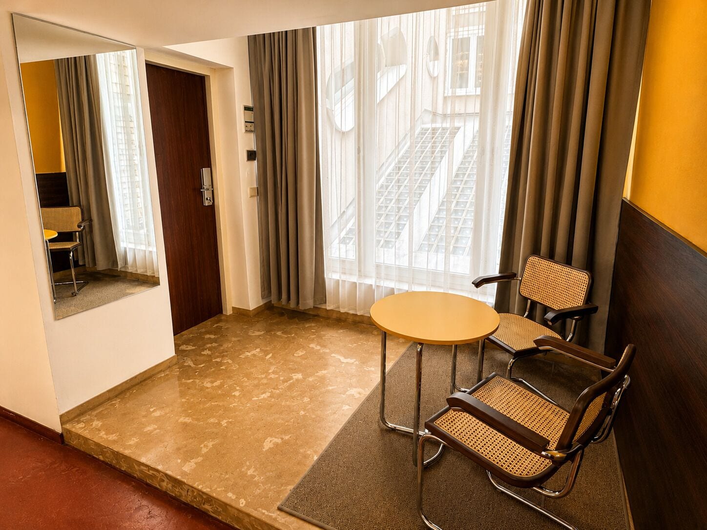 Habitación de hotel con una pequeña mesa redonda, dos sillas, un espejo y vistas a un edificio a través de cortinas translúcidas.
