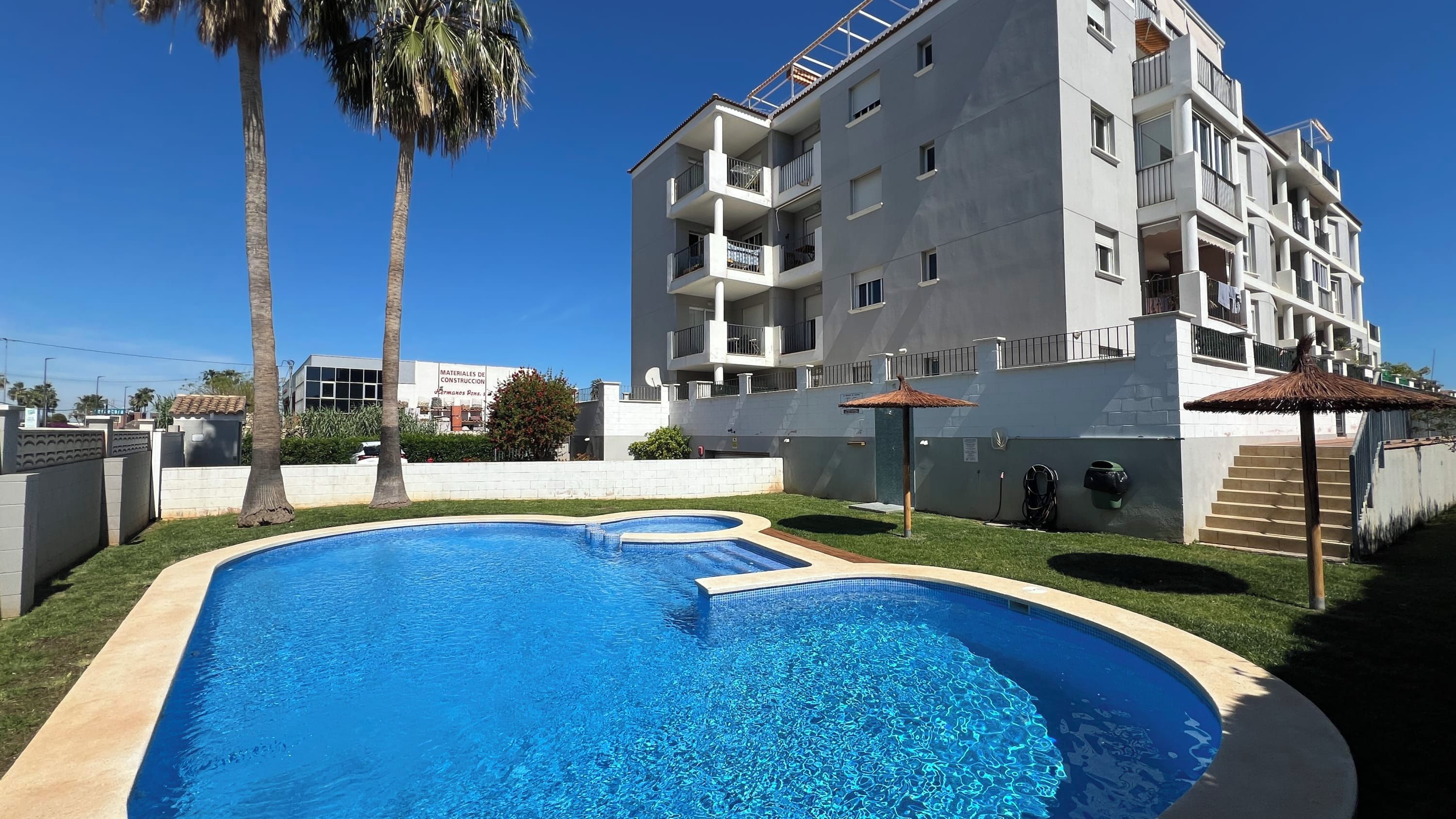 A swimming pool with a clear blue water, surrounded by a white building with balconies and a palm tree.