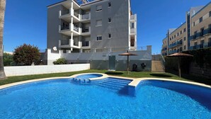 Una piscina d'aigua cristal·lina i blava, envoltada d'un edifici blanc amb balcons i una petita zona de jardí amb una ombra de palla.