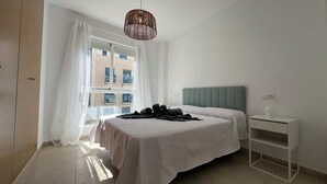 Un dormitori amb un llit, una taula de nit, una làmpada penjant, una finestra amb cortines i vista a edificis a l'exterior.