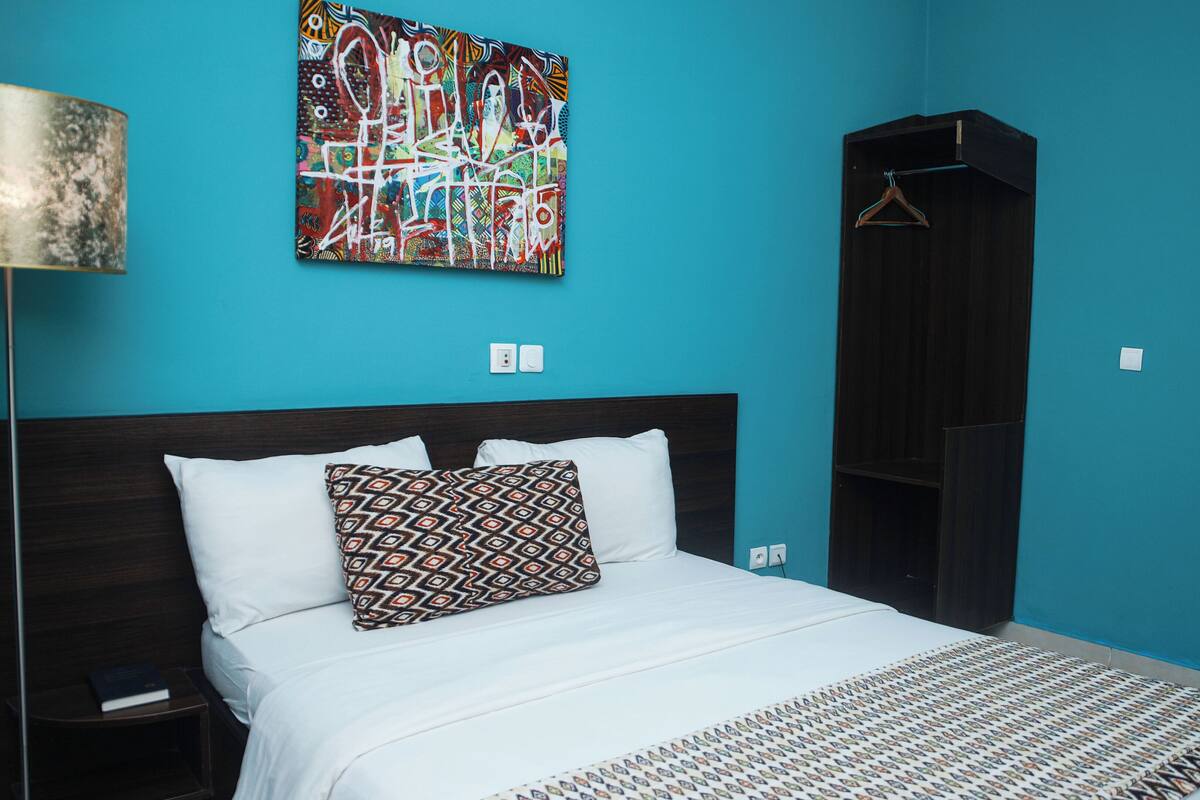Una cama bien tendida con almohadas estampadas, un cabecero oscuro y un cuadro abstracto de colores en la pared.