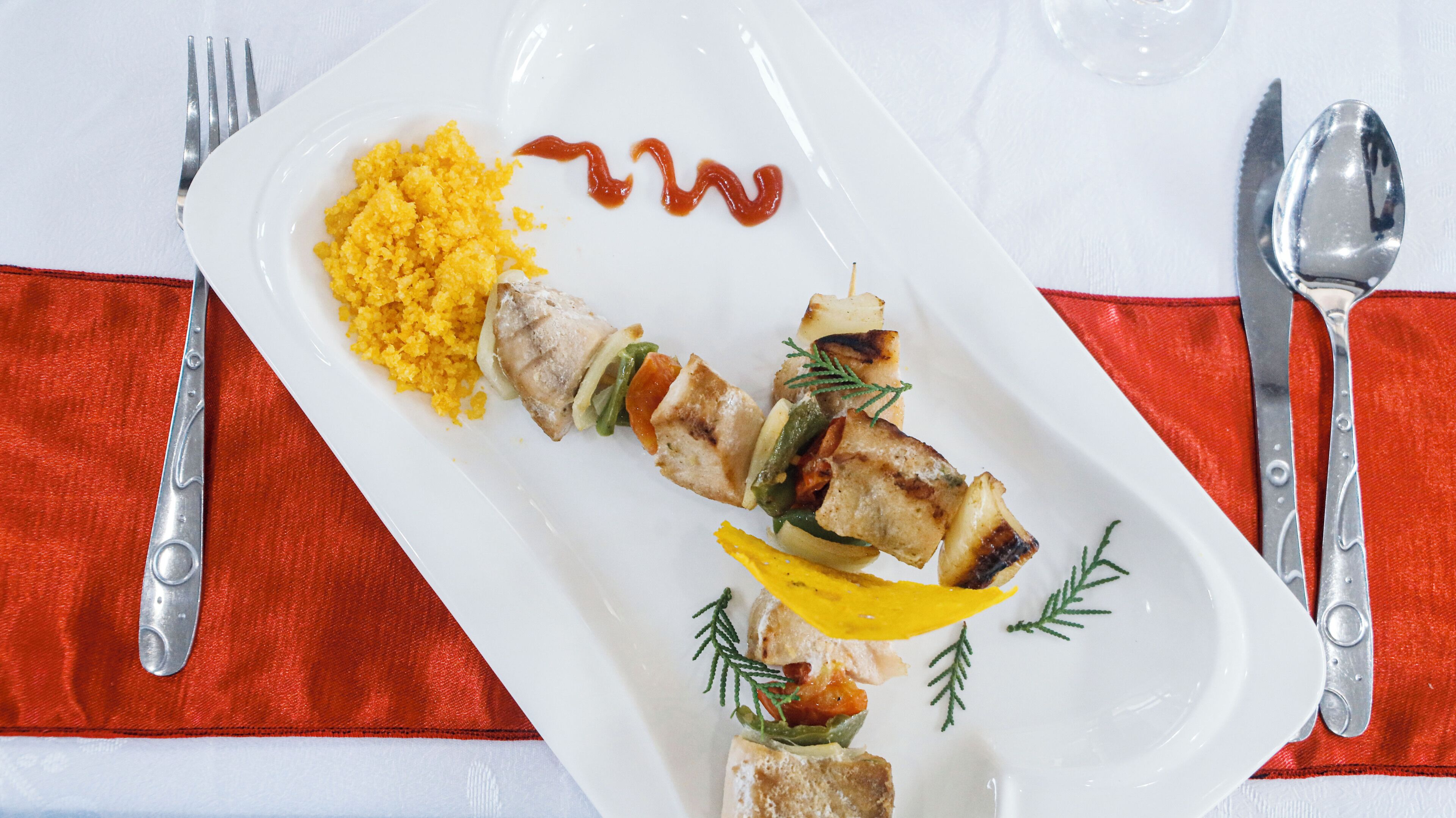 A white rectangular plate with grilled meat, vegetables, and yellow rice, accompanied by a red sauce and a spoon.