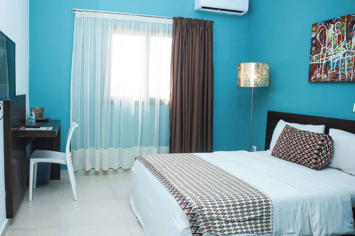 Habitación de hotel con cama, escritorio con silla, televisión, ventana con cortinas y un cuadro en la pared.