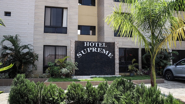 A hotel sign with the name "HOTEL SUPREME" and a modern building facade with multiple windows and a well-maintained garden area.