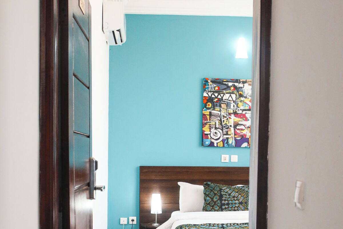 Habitación de hotel con cama, mesita de noche, lámpara y un cuadro abstracto de colores en la pared.